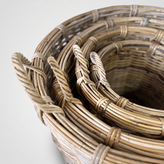 A close-up of a coiled natural rattan basket or rug with woven details and twisted loop handles, photographed on a light neutral background.