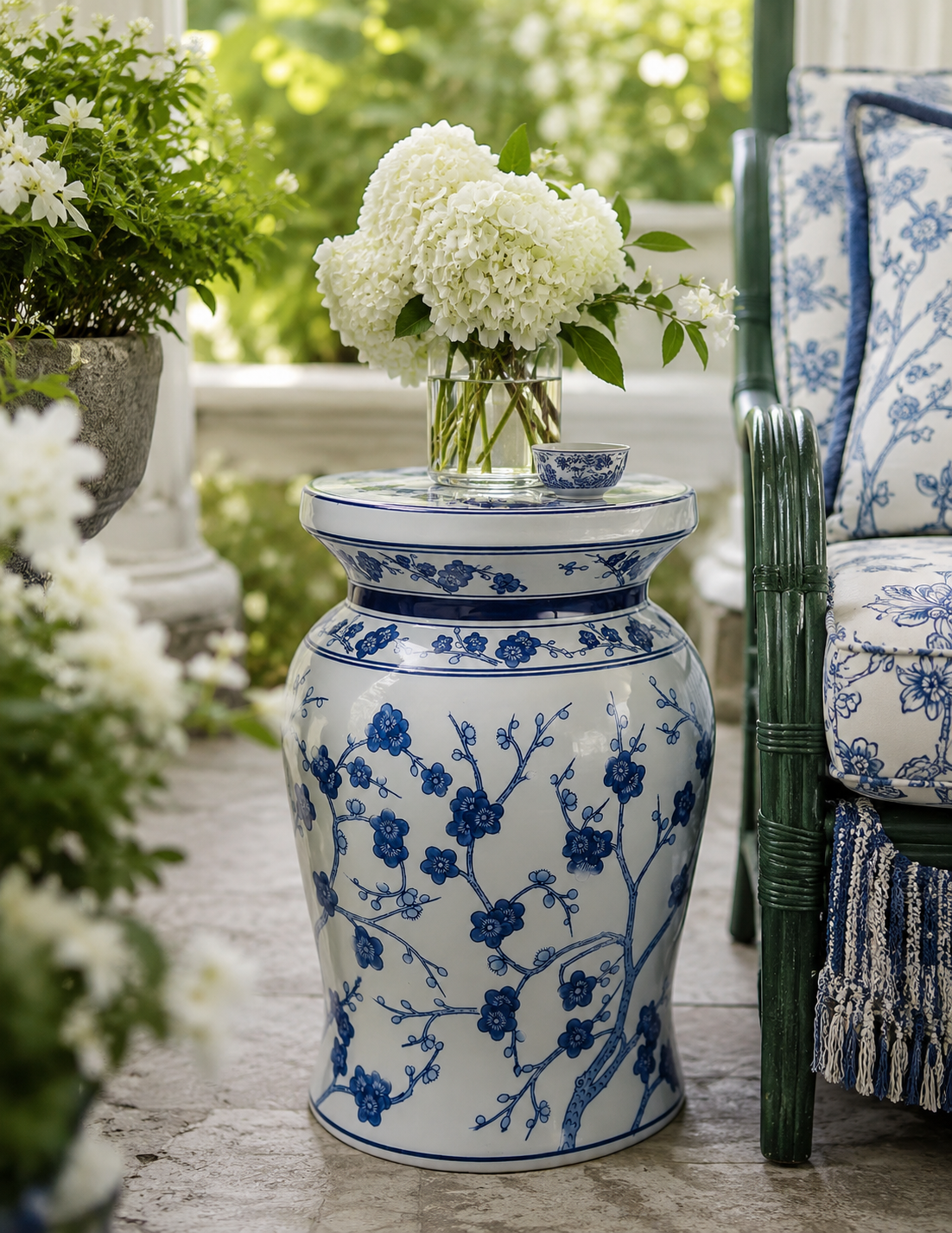 A blue and white porcelain garden stool with floral branch patterns, styled on a stone patio with white flowers on top and a matching blue-and-white cushioned chair beside it.