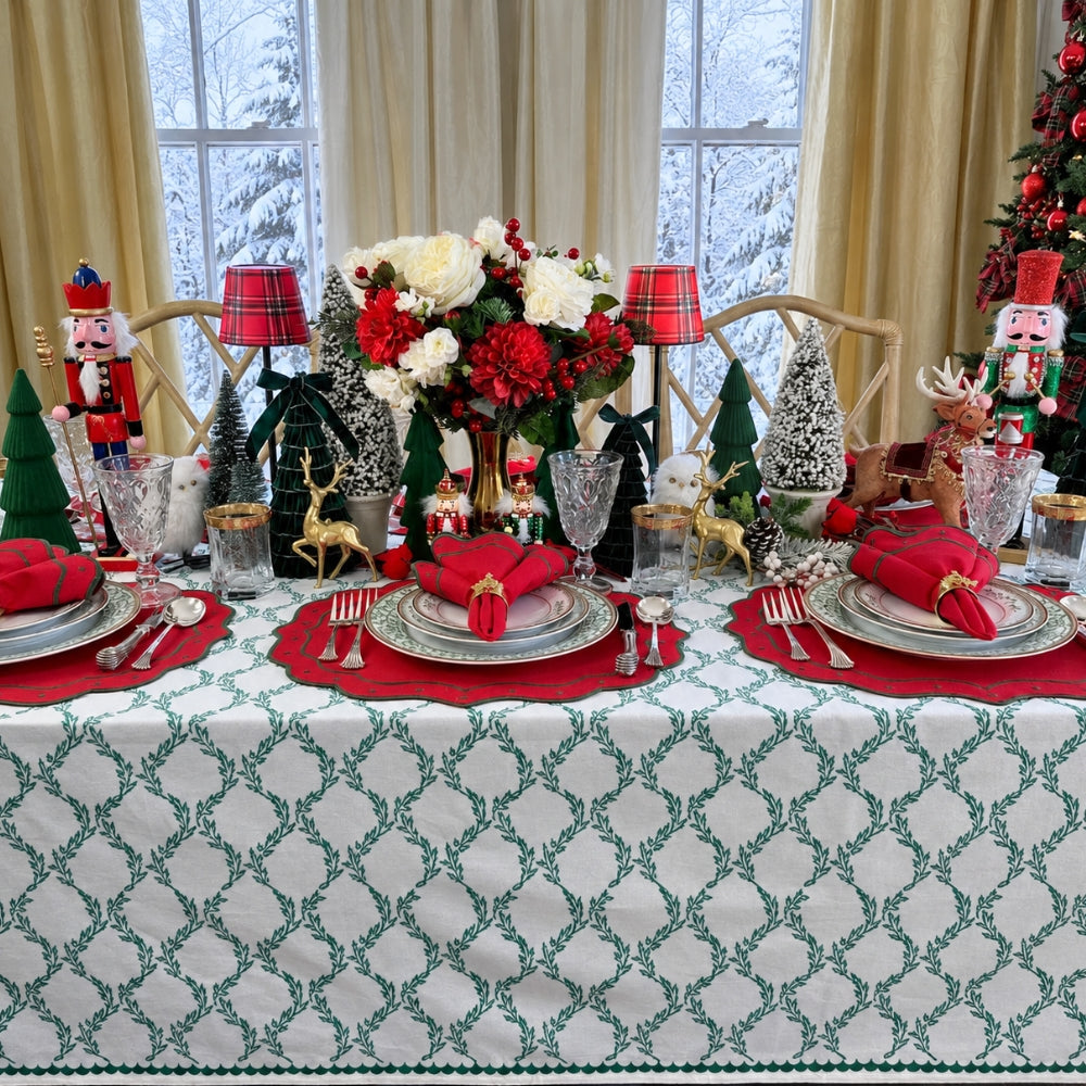 A festive holiday table setting decorated with a green and white patterned tablecloth, red placemats, silver plates, red napkins, a floral centerpiece, and Christmas ornaments including nutcrackers, mini trees, and reindeer figurines.