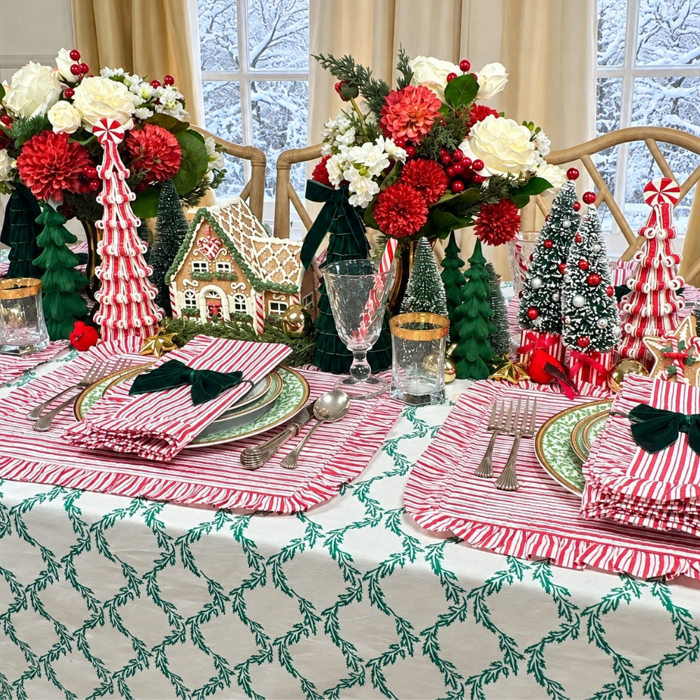 A festive Christmas table setting featuring red and white striped placemats with green leafy trim, matching folded napkins with black bow accents, a white tablecloth with a green geometric pattern, and multiple holiday floral arrangements with red and white flowers, miniature trees, and a gingerbread house centerpiece.
