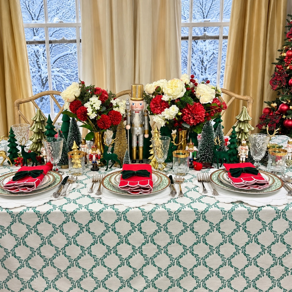 A beautifully set holiday table with a green and white patterned tablecloth, place settings with red napkins, and festive centerpieces featuring red and white flowers, miniature trees, nutcrackers, and gold ornaments in front of large windows with beige curtains.