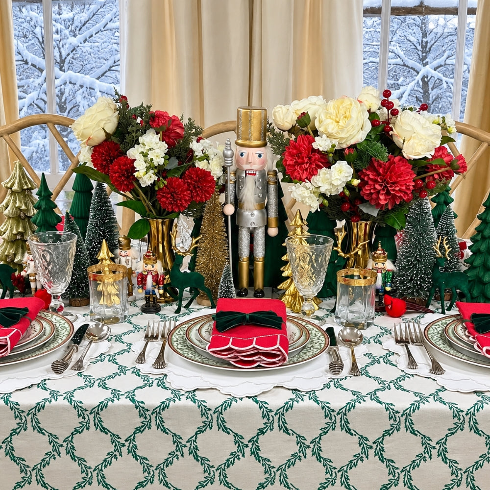 A festive holiday table setting decorated with red and white floral arrangements, miniature green Christmas trees, gold nutcrackers, deer figurines, and matching place settings with red napkins and black bows. The table is covered with a white tablecloth featuring a green leaf pattern, set against a background of beige curtains and snowy window views.