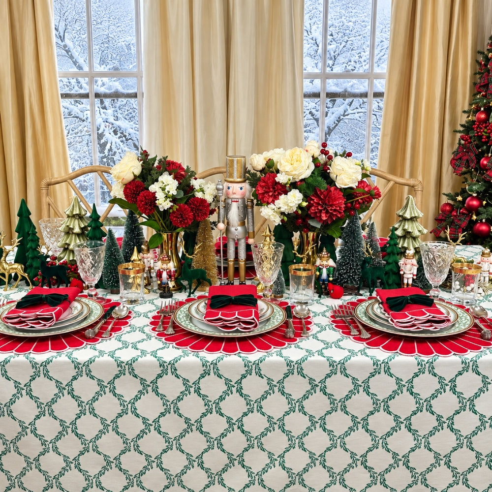A festive Christmas table setting with a green and white patterned tablecloth, red and black napkins, silver plates, crystal glasses, floral centerpieces, nutcracker decorations, and a Christmas tree in the background.