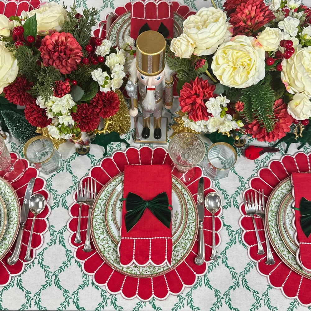 A festive holiday table setting featuring red scalloped placemats, matching red napkins with black bow accents, green and white patterned plates, silver cutlery, and large floral centerpieces with red and cream flowers.