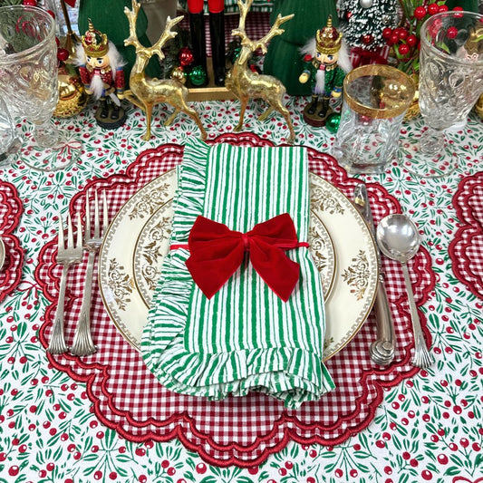 A festive Christmas table setting featuring a green and white striped napkin tied with a red velvet bow, layered over a white and gold charger plate on a red scalloped placemat. The tablecloth has a red and white gingham pattern with green berries, surrounded by silver cutlery, crystal glasses, and holiday decorations in the background.