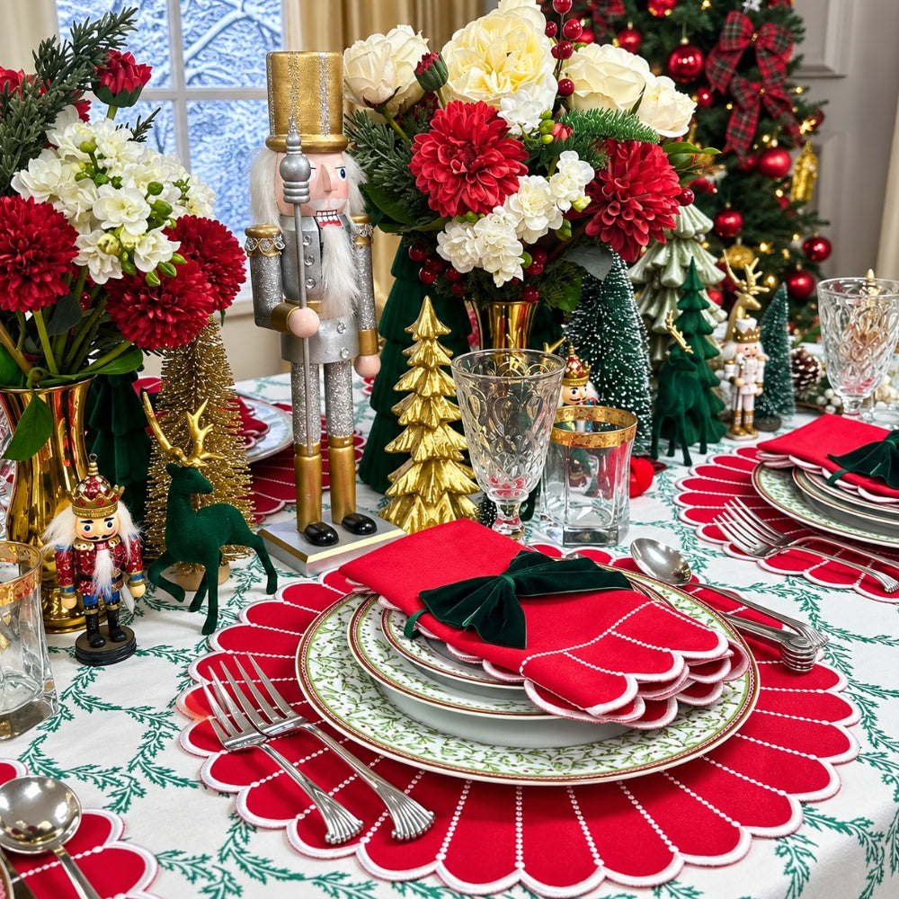 A festive Christmas table setting with red and white scalloped placemats, matching napkins tied with dark green velvet bows, decorative plates, glassware, nutcrackers, green trees, and a floral centerpiece in front of a decorated tree.