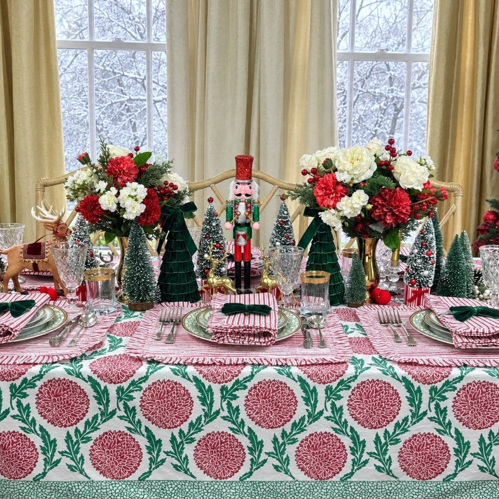 A festive holiday table setting decorated with a pink and green patterned tablecloth, matching napkins, small Christmas trees, floral arrangements, and a nutcracker centerpiece in front of large windows with cream curtains.