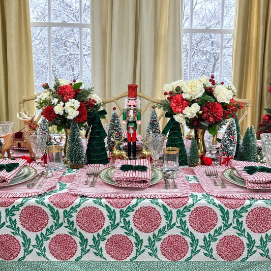 A festive holiday table setting decorated with a pink and green patterned tablecloth, matching napkins, small Christmas trees, floral arrangements, and a nutcracker centerpiece in front of large windows with cream curtains.