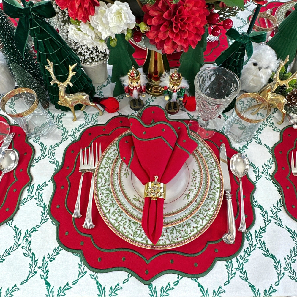 A festive Christmas table setting featuring a red scalloped placemat, a white and green patterned dinner plate, a folded red napkin with a gold-toned ring, silver cutlery, and holiday decorations including green trees, red flowers, and nutcrackers.