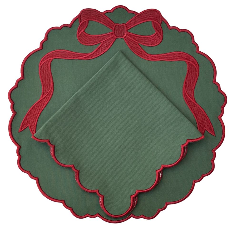 A green square cloth napkin folded neatly inside a scalloped round green napkin holder or placemat with a red embroidered bow at the top and red scalloped trim.