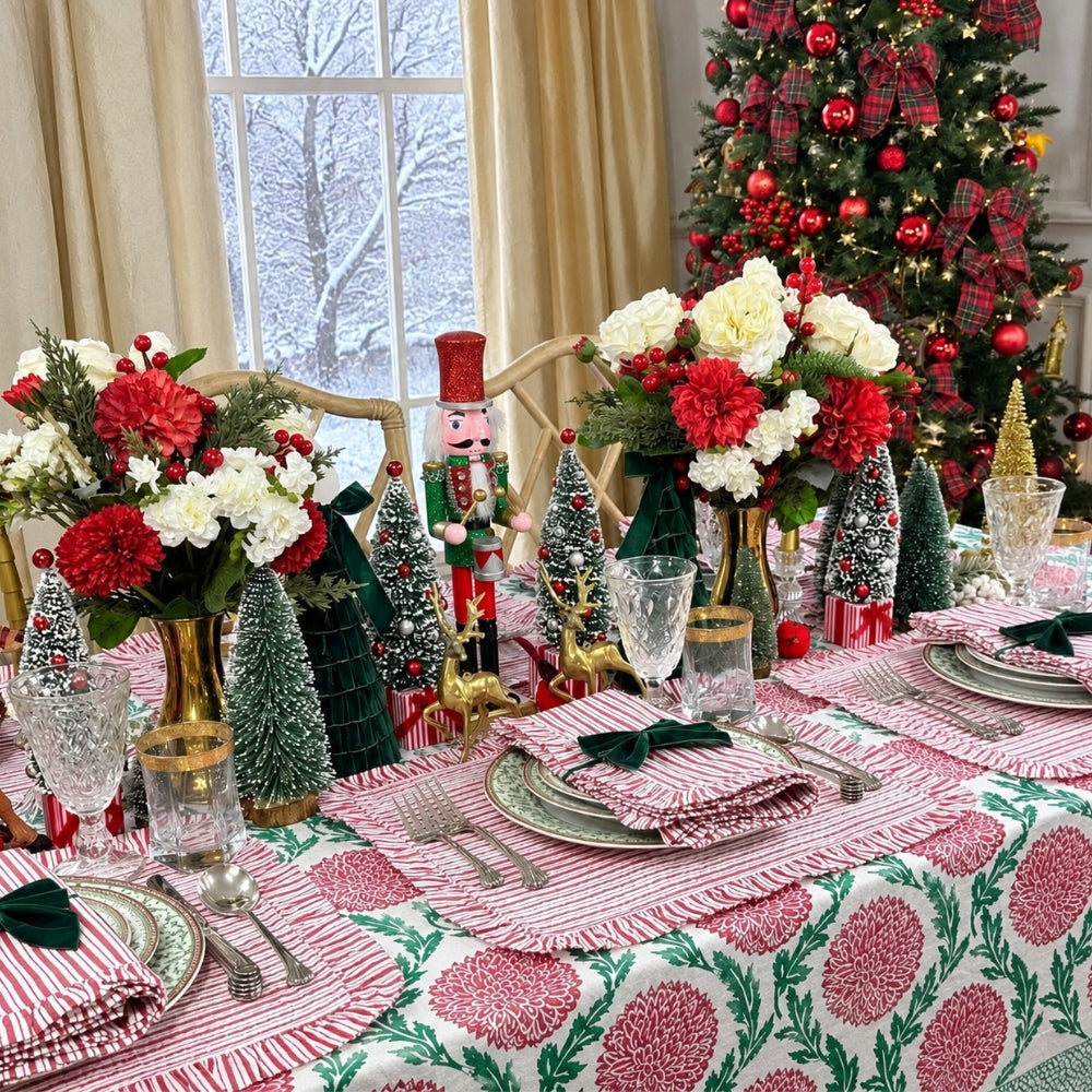 A festive Christmas table setting with red and white striped placemats, matching napkins with green bows, floral centerpieces, miniature Christmas trees, and a decorated Christmas tree in the background.