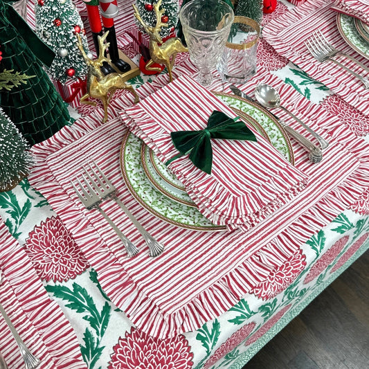 A festive holiday table setting featuring a red and white striped tablecloth with green floral patterns, matching napkins tied with a green bow, green and gold patterned plates, silver cutlery, and Christmas tree decorations.