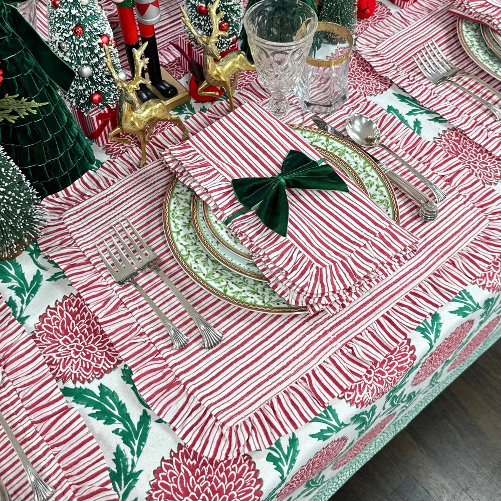 A festive holiday table setting featuring a red and white striped tablecloth with green floral patterns, matching napkins tied with a green bow, green and gold patterned plates, silver cutlery, and Christmas tree decorations.