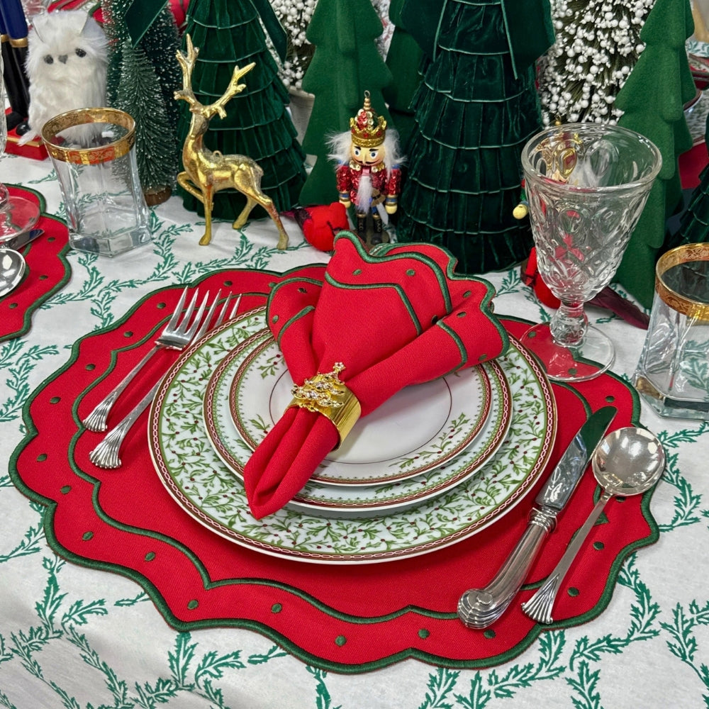 A festive Christmas table setting featuring a red scalloped placemat with green leaf trim, layered white and gold dinner plates, a red napkin with green trim, silver cutlery, and holiday decorations in the background.