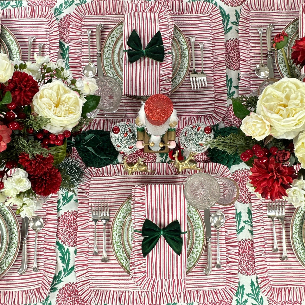 A festive holiday table setting featuring red and white striped napkins with dark green bow accents, silver flatware, patterned plates, and floral arrangements with red and white roses, green foliage, and nutcracker decorations.