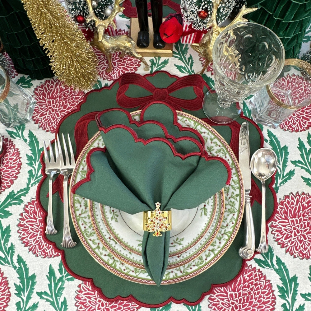 A festive holiday table setting featuring a green and red scalloped placemat, white patterned dinner plates, a dark green napkin with scalloped edges, a gold-toned napkin ring, silver cutlery, and a red-and-white floral tablecloth.