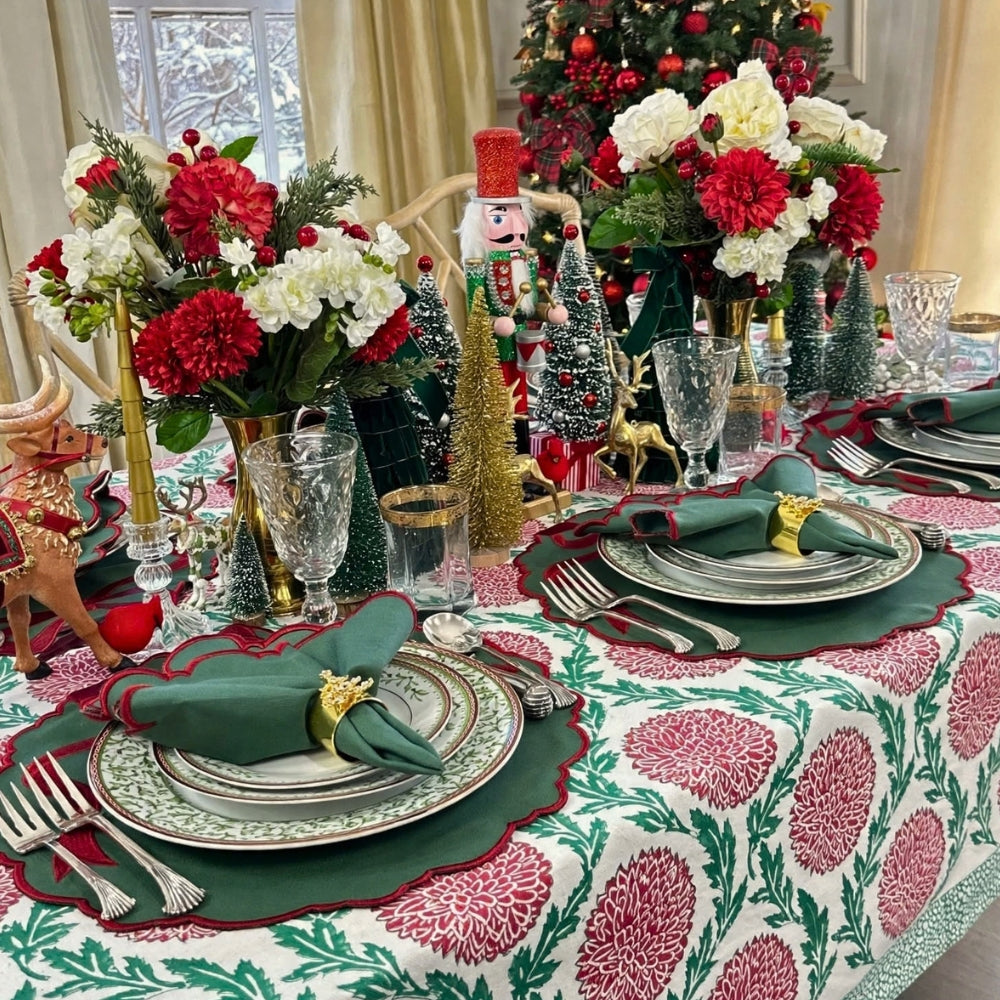 A festive Christmas table setting with a white tablecloth printed with red and green floral motifs, green napkins with gold rings, clear glassware, and holiday decorations including miniature trees, a nutcracker, and floral centerpieces.