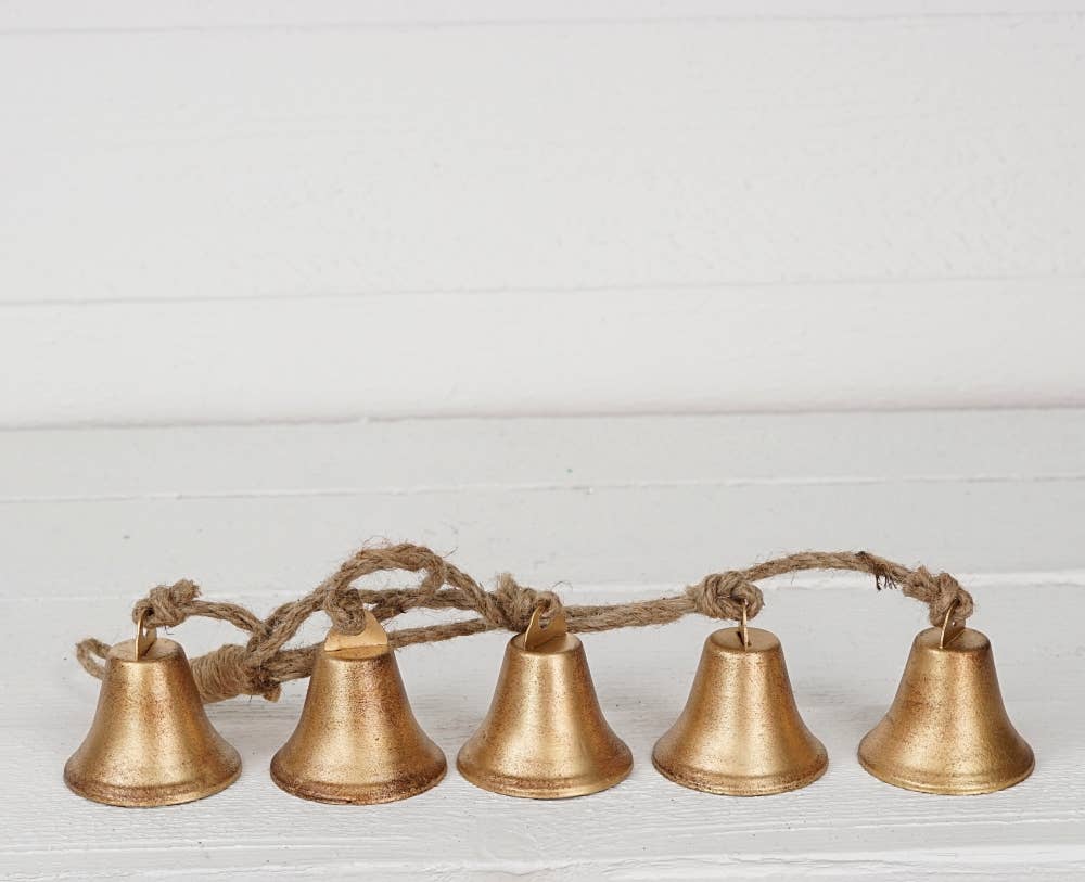 Five small metallic gold bells strung together with a natural jute rope, displayed against a white wood paneled background.
