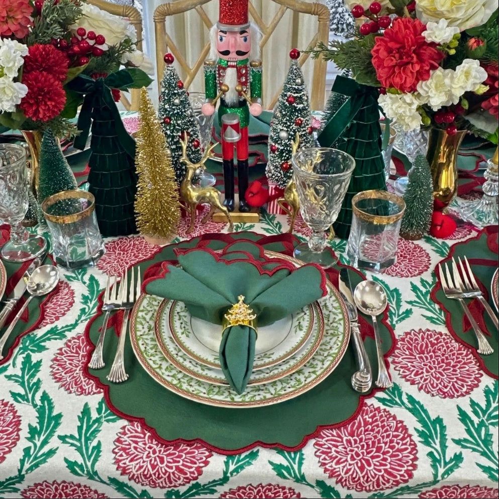 A festive Christmas table setting featuring a green and red patterned tablecloth, matching napkins, layered plates, gold flatware, and decorative holiday centerpieces with miniature trees, a nutcracker, and floral arrangements.