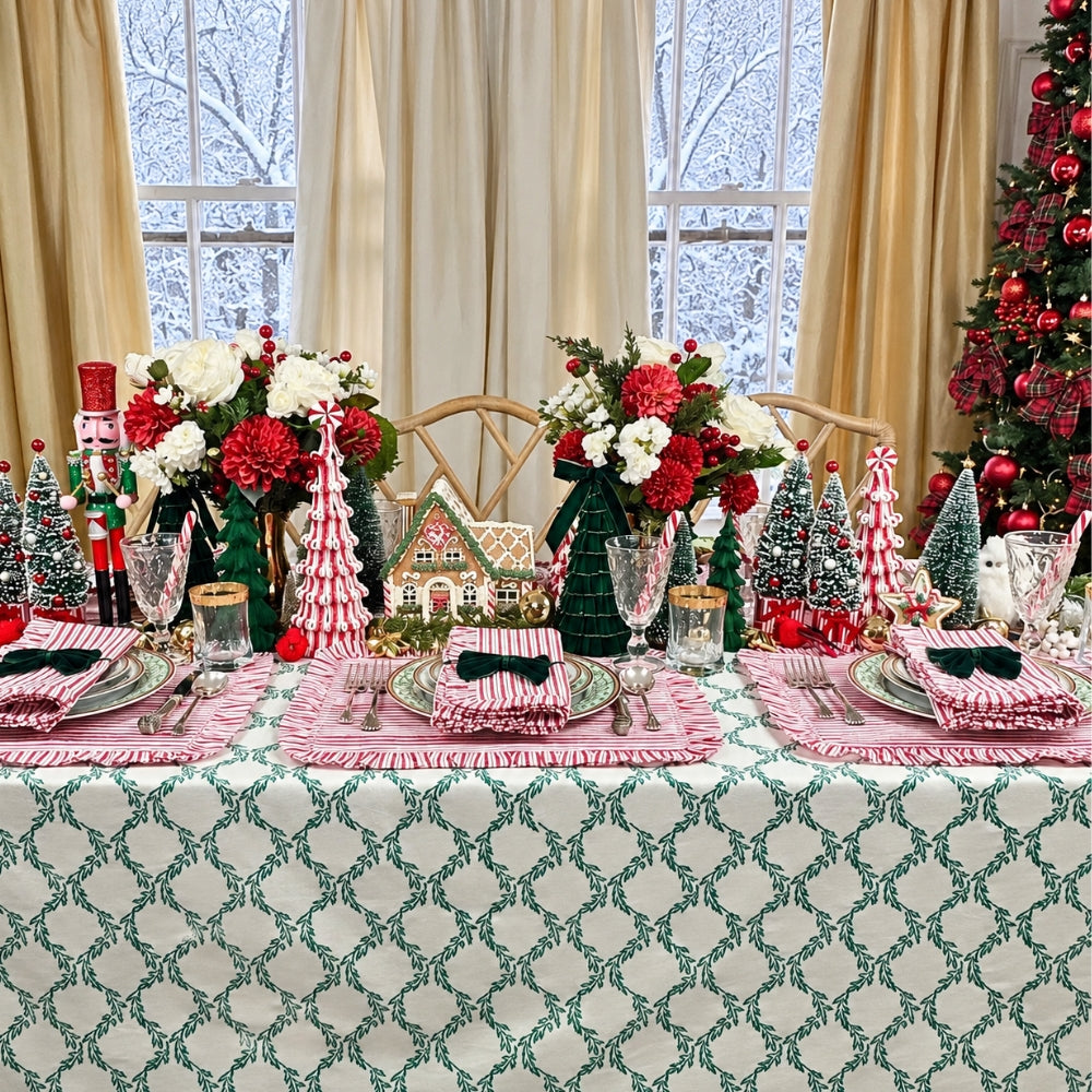 A festive Christmas table setting with a green and white patterned tablecloth, pink and white striped napkins, glassware, floral centerpieces, miniature Christmas trees, gingerbread houses, and nutcracker decorations in front of large windows with beige curtains.