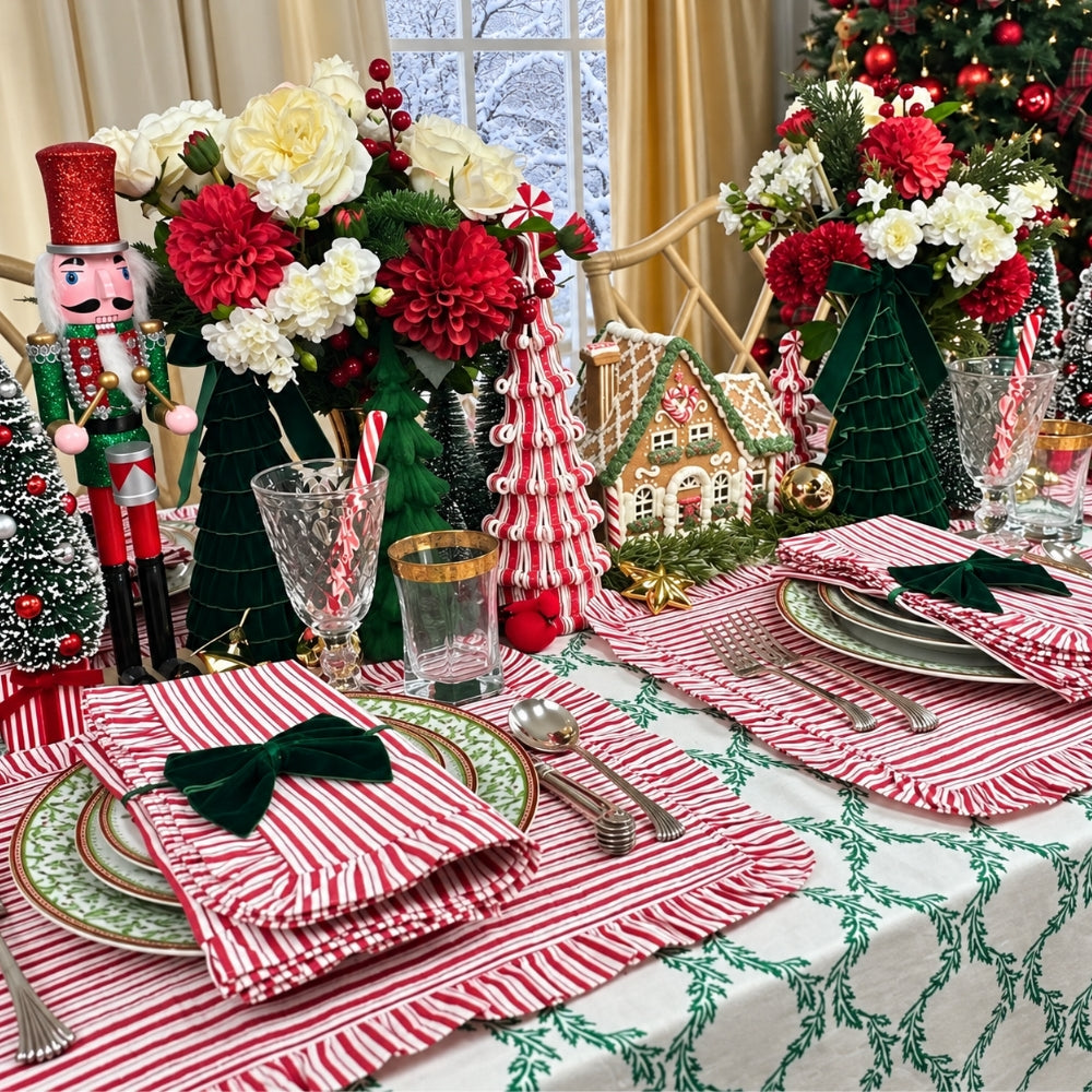 A festive Christmas table setting with red and white striped napkins tied with dark green bows, matching striped placemats, green and white patterned plates, clear glasses with gold trim, floral arrangements, a nutcracker, gingerbread house decor, and a Christmas tree in the background.