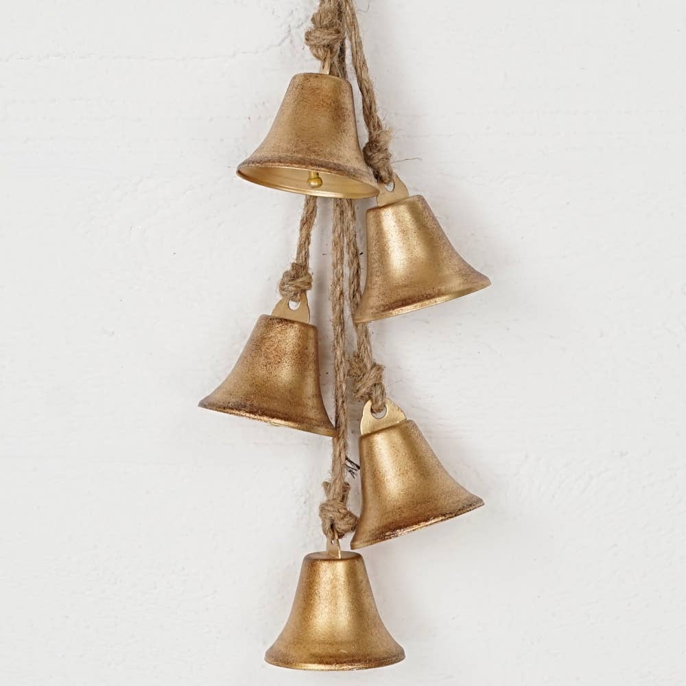 A decorative hanging cluster of five small metallic bells in a distressed gold finish, suspended from a natural jute rope against a white textured wall.