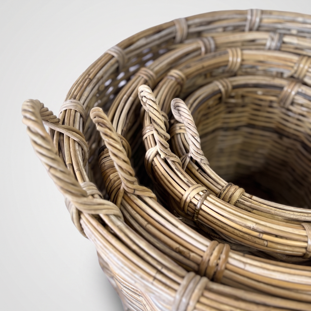A close-up of a coiled natural rattan basket or rug with woven details and twisted loop handles, photographed on a light neutral background.