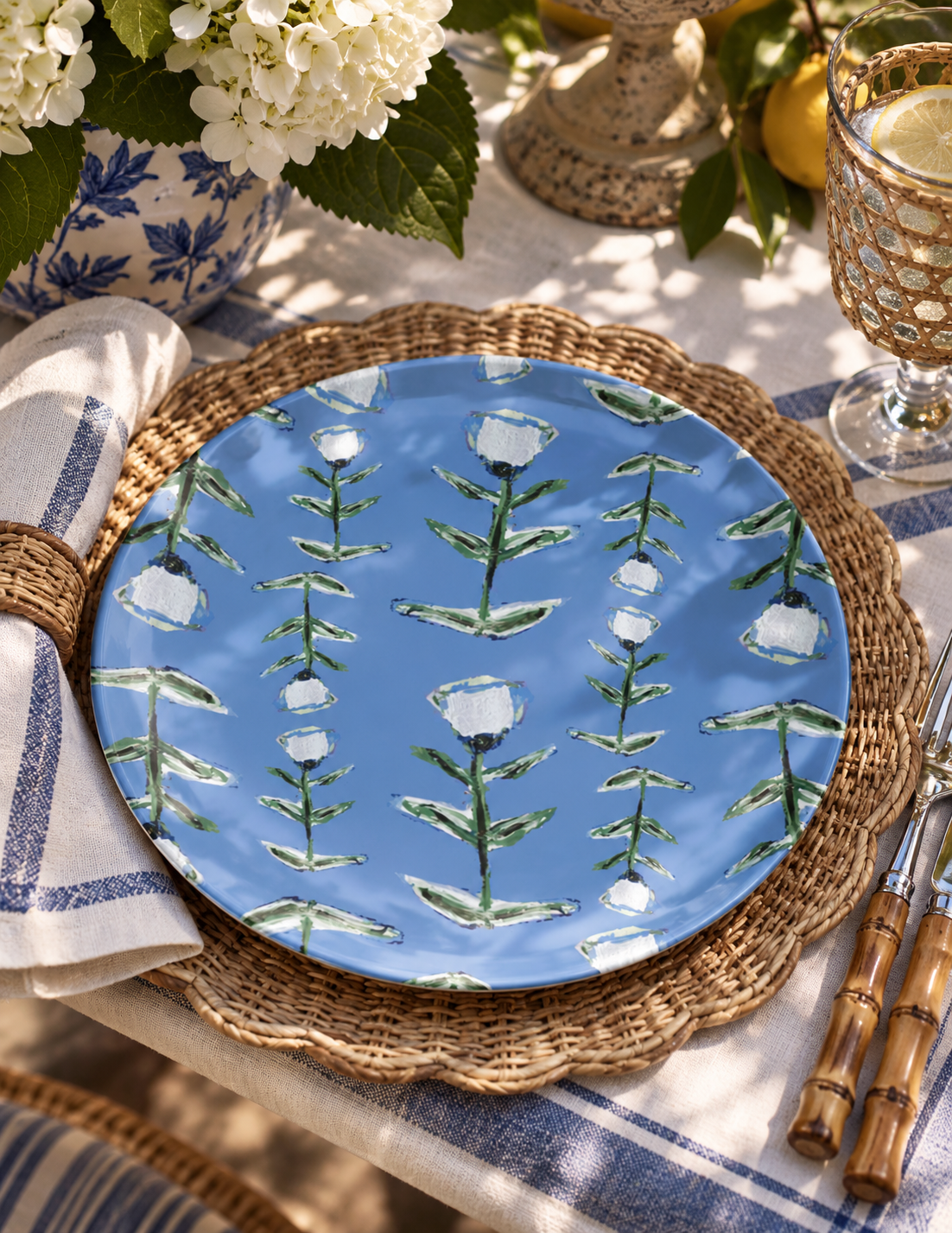 A round blue ceramic plate with a green and white floral vine pattern, styled on a woven placemat over a blue-and-white striped napkin in an outdoor dining setting.