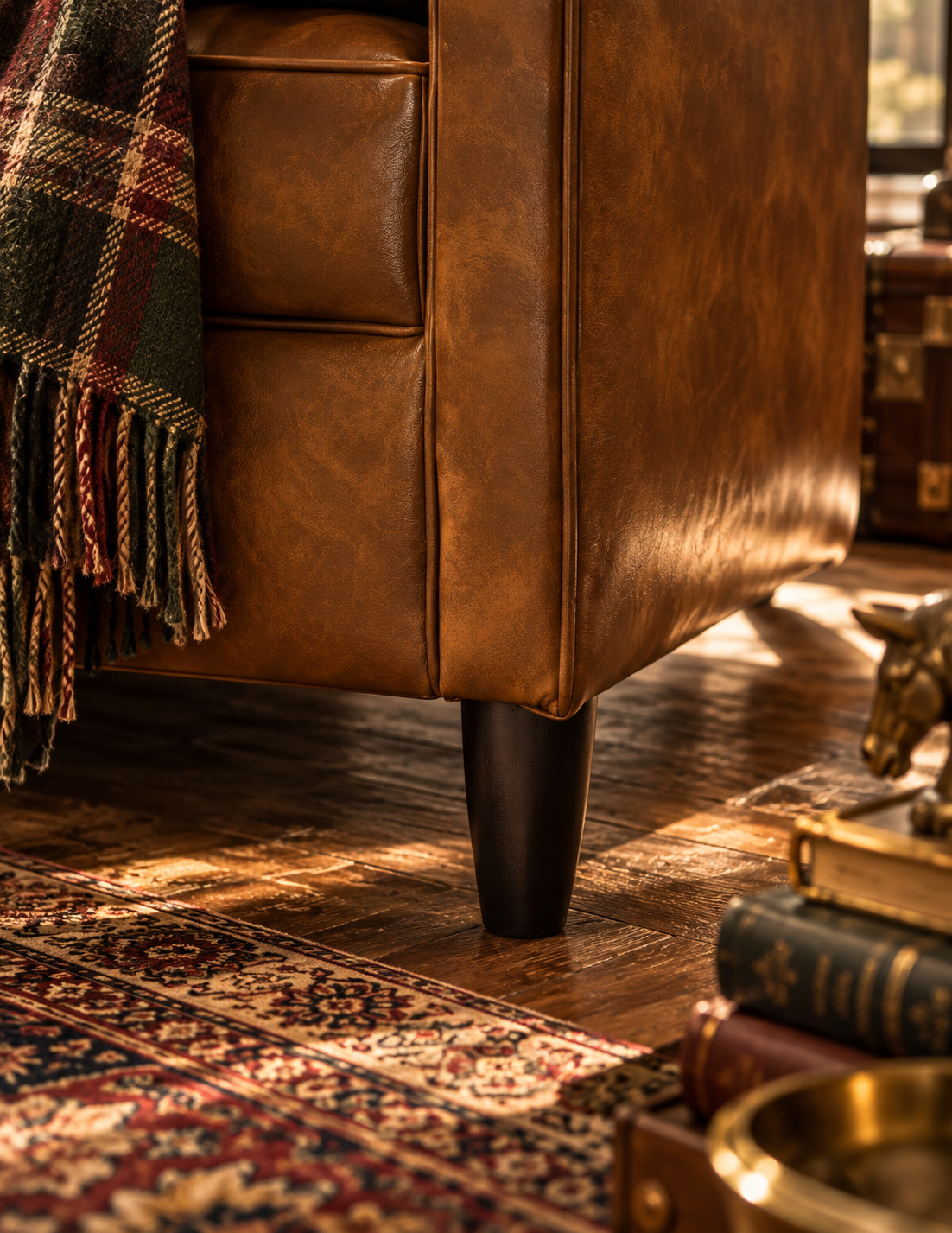 A close-up of a brown leather armchair in a warm, sunlit room, with a plaid throw blanket draped over the side and a dark wooden floor beneath.