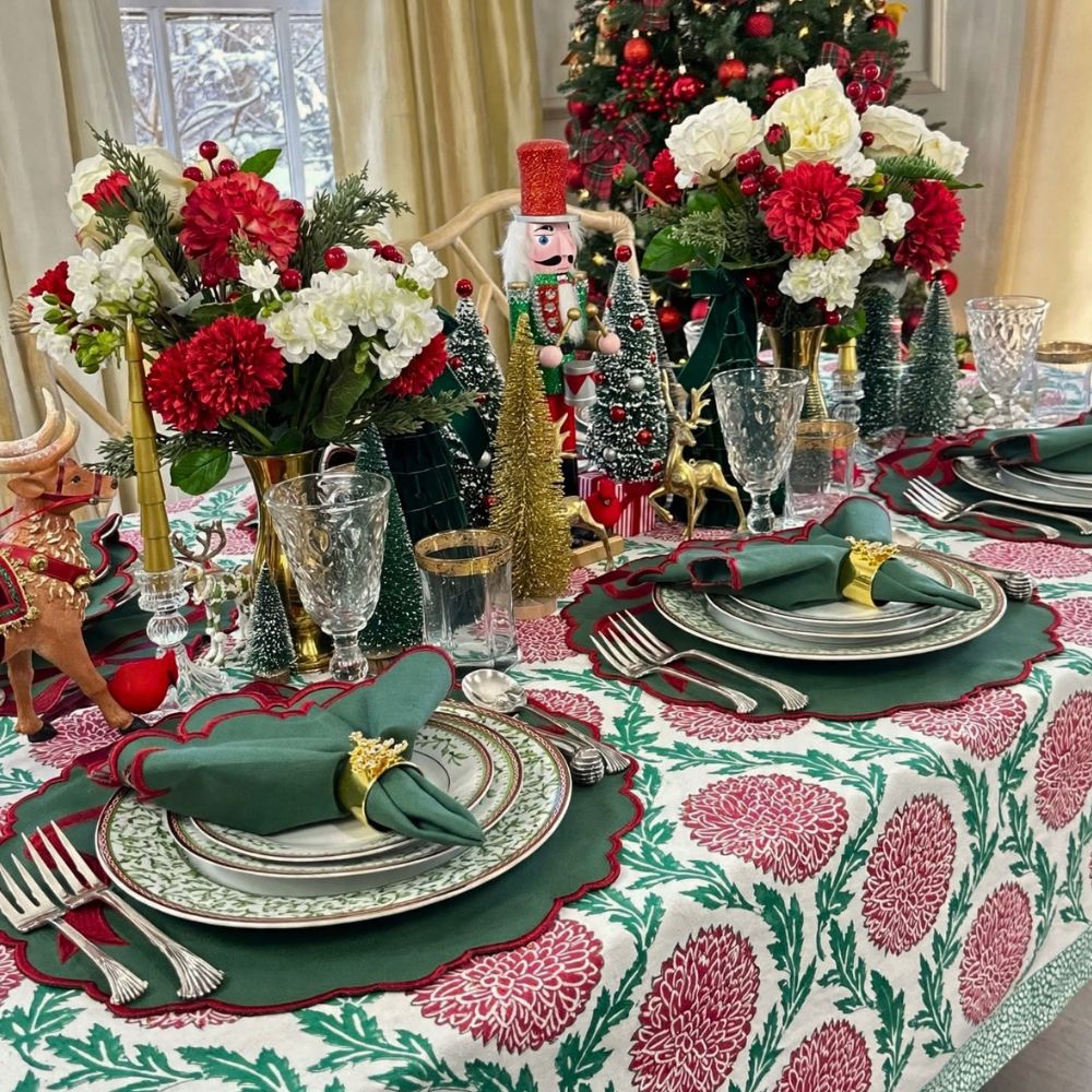 A festive Christmas table setting with a red and green floral tablecloth, matching scalloped placemats, green napkins with gold rings, and holiday decorations including miniature trees, a nutcracker, and floral arrangements.