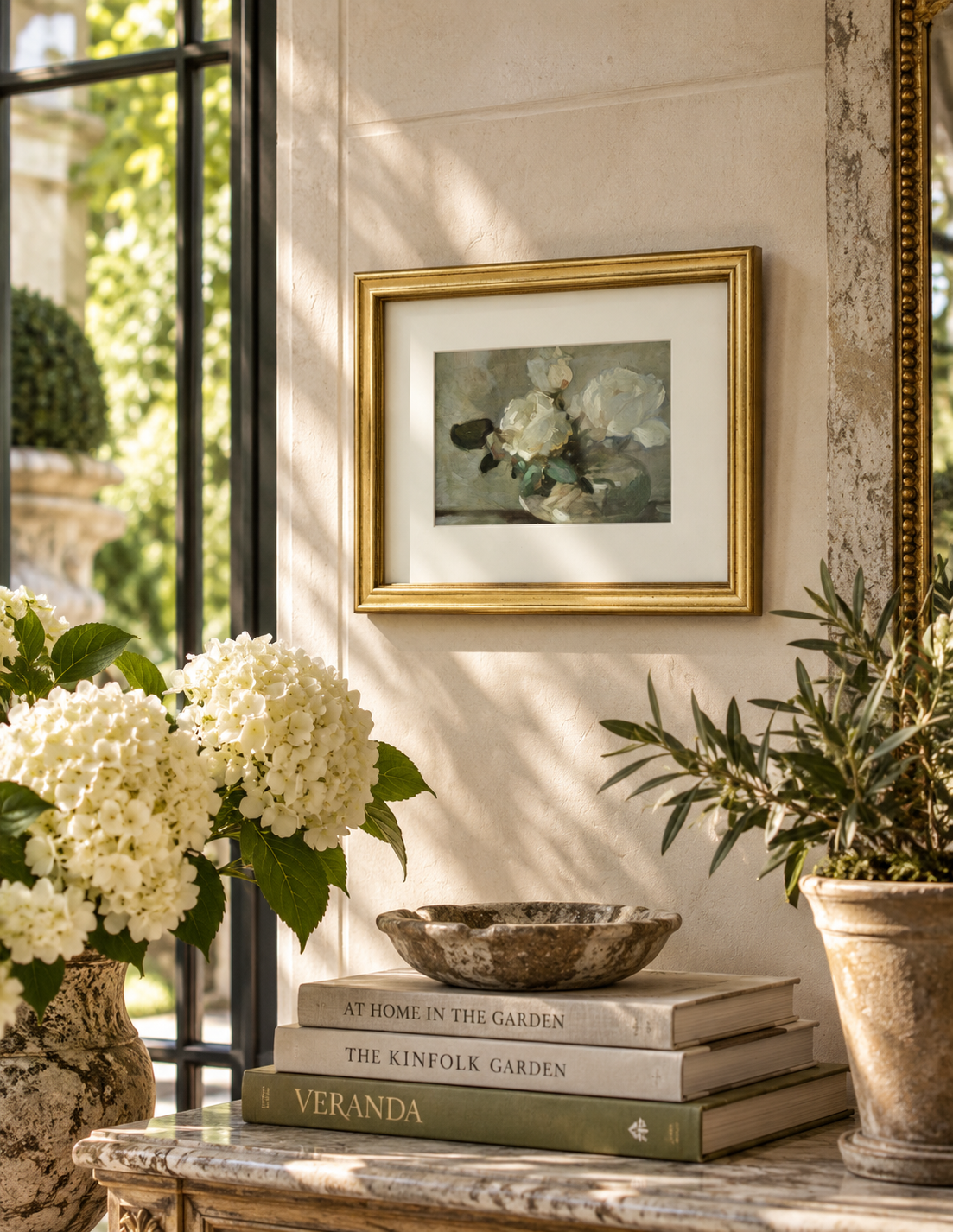 A framed floral art print with white flowers and green leaves, set in a gold frame with a white mat, hanging on a beige wall in a styled interior scene with books, a bowl, and potted plants.
