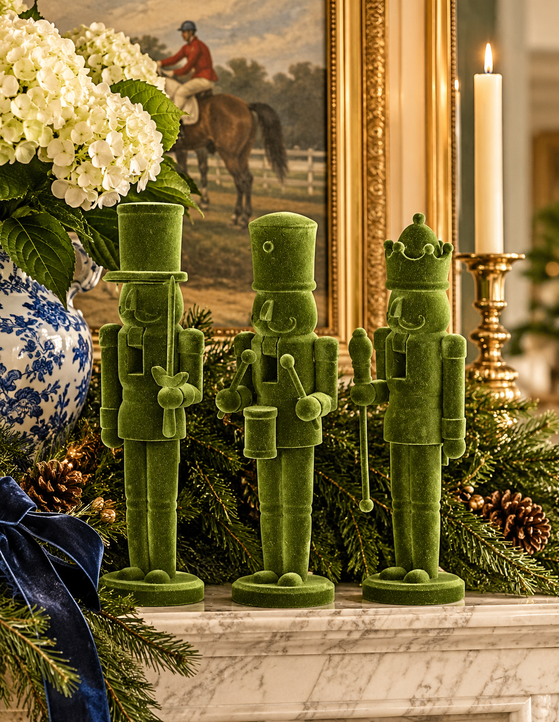 Three green velvet nutcracker figurines displayed on a marble mantel in a decorated holiday setting with pine branches, a blue-and-white ceramic vase of white flowers, a lit candle, and a framed painting in the background.