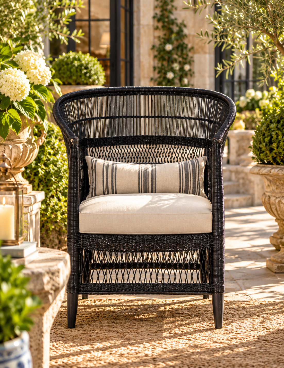 A black woven outdoor armchair with a cream seat cushion and a striped accent pillow, set on a stone patio in a garden courtyard.