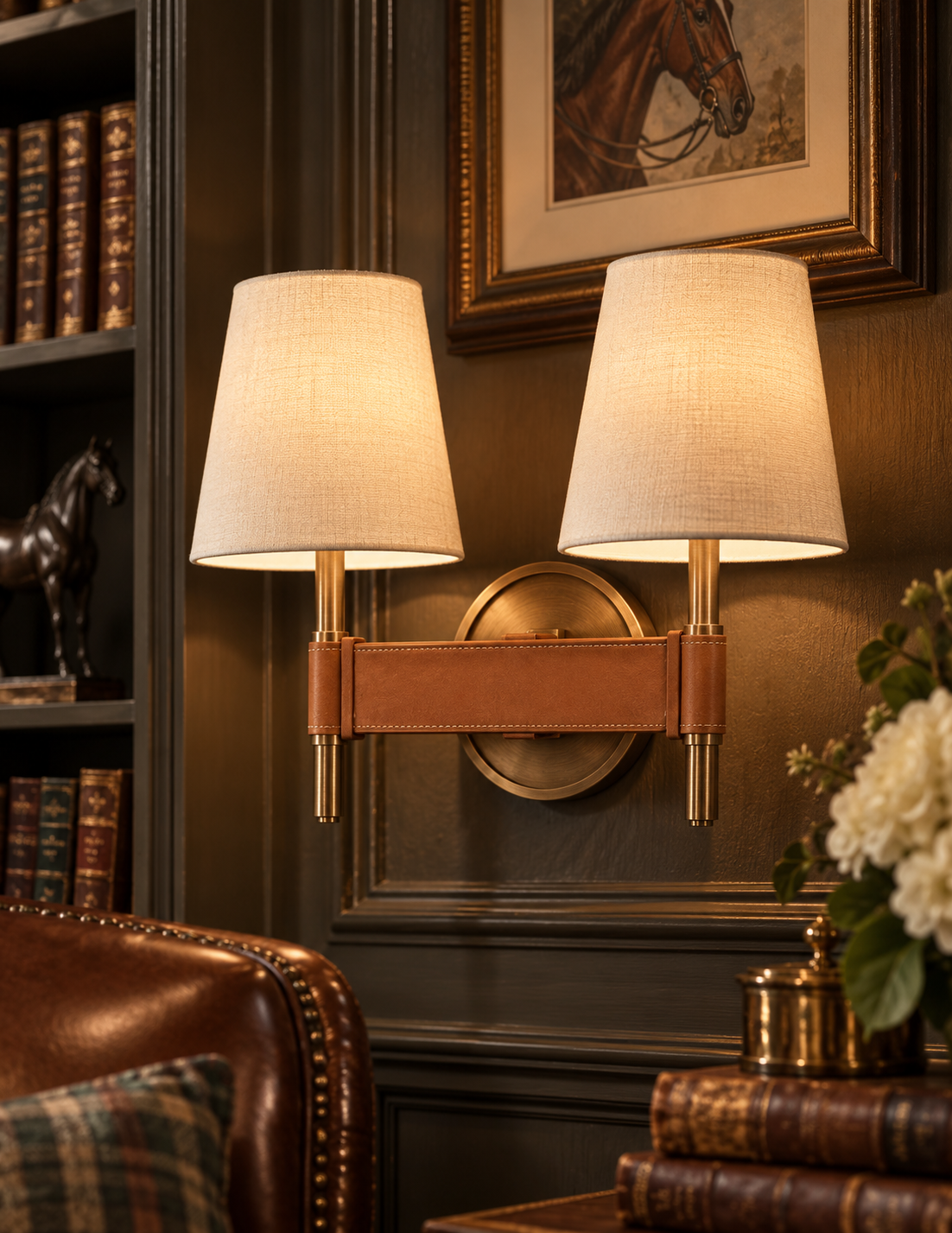 A warm brass double wall sconce with two beige linen drum shades and a tan leather strap detail, mounted on a dark wood paneled wall in a classic library setting.