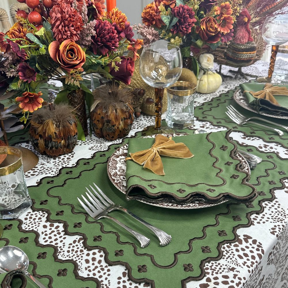 An autumn-themed table setting featuring a green embroidered table runner and matching napkins tied with gold ribbon, layered over a white lace tablecloth. The setting includes fall floral arrangements, decorative pumpkins, and silver flatware.