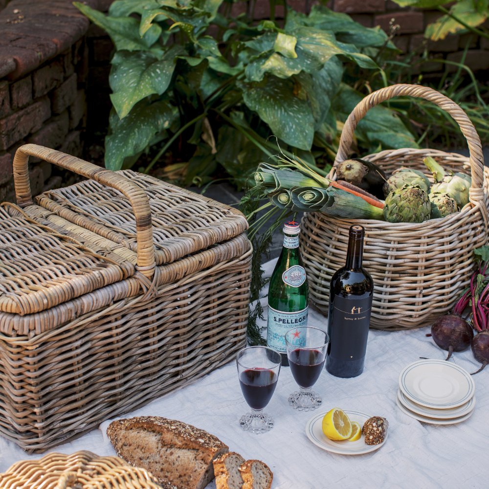 A styled picnic setup outdoors on a white tablecloth with two wicker picnic baskets, bottles of wine, two glasses of red wine, bread, lemons, and vegetables, with green foliage and a brick wall in the background.