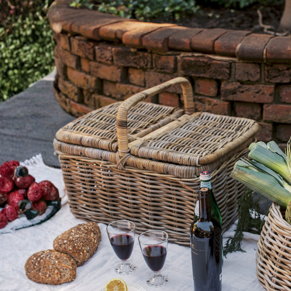 A large woven wicker picnic basket with a hinged lid sits on a white tablecloth outdoors beside two filled wine glasses, a bottle of red wine, bread, and fresh fruit. A brick fire pit is visible in the background.