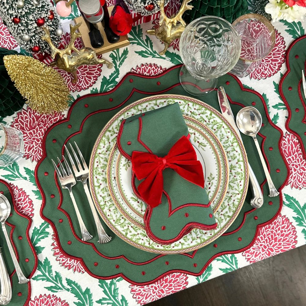A festive Christmas table setting featuring a green and red scalloped placemat, white patterned plates, silver cutlery, a green napkin with a red bow, and a matching patterned tablecloth.