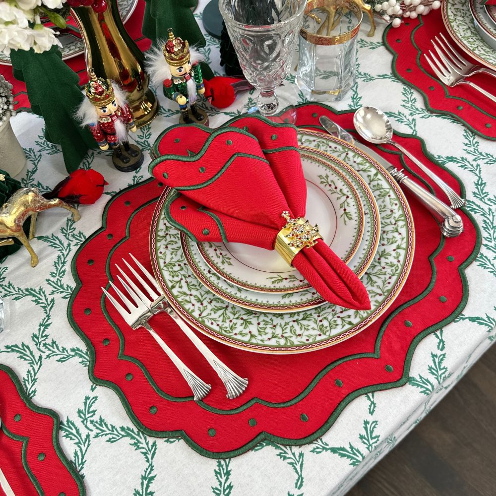 A festive holiday table setting featuring red scalloped table placemats with green leaf patterns, white plates with gold trim, a red napkin, and silver cutlery, surrounded by Christmas decorations.