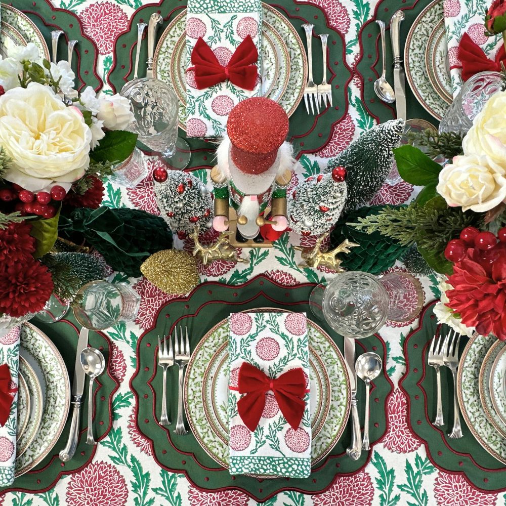 A festive Christmas table setting featuring coordinated place settings with green and red floral patterns, white roses, red berries, clear glass goblets, silver cutlery, and a small nutcracker centerpiece.