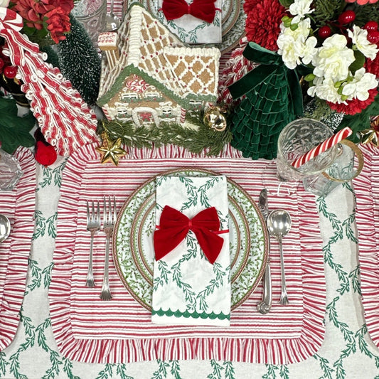 A festive holiday table setting featuring red and white striped placemats with fringe, matching napkins tied with red bows, silver cutlery, and Christmas decorations including a gingerbread house, green velvet tree ornaments, and red-and-white candy canes.