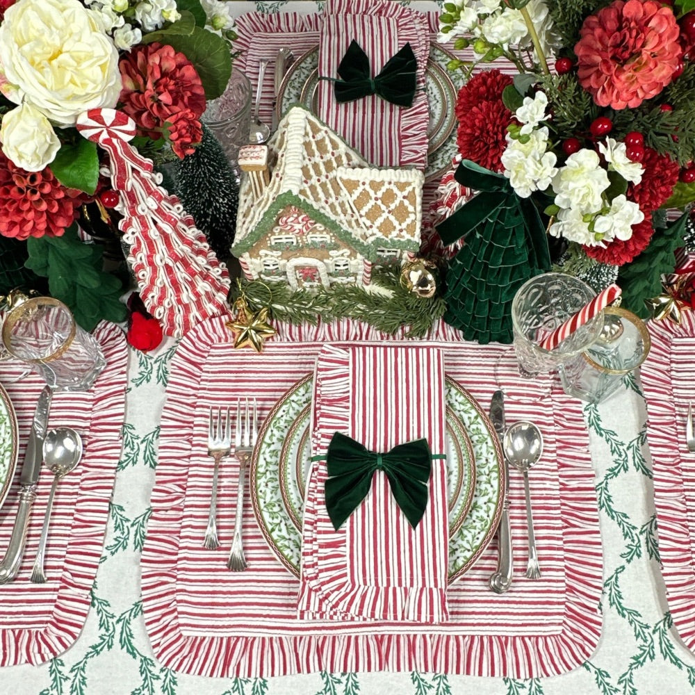 A festive Christmas table setting featuring red and white striped placemats with green leaf patterns, matching green bow napkins, silver flatware, clear drinking glasses, and floral centerpieces with red and white flowers.