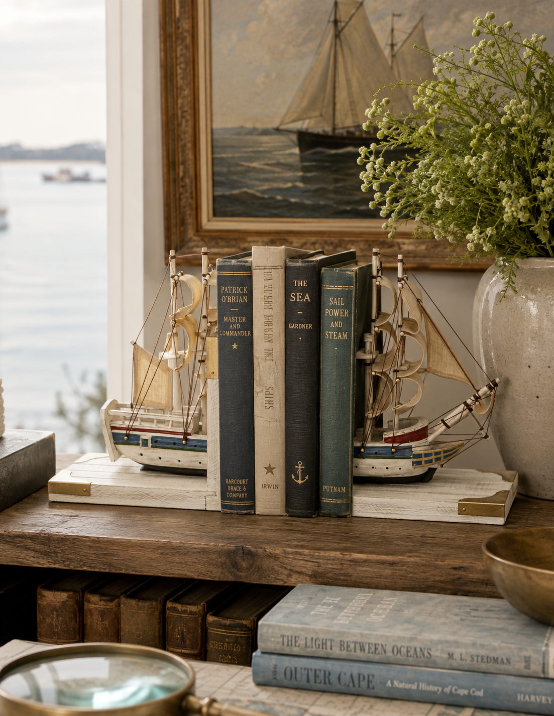A pair of decorative model sailing ships bookends holding a set of vintage nautical books on a wooden console table, with a framed ocean painting and a potted plant in the background.