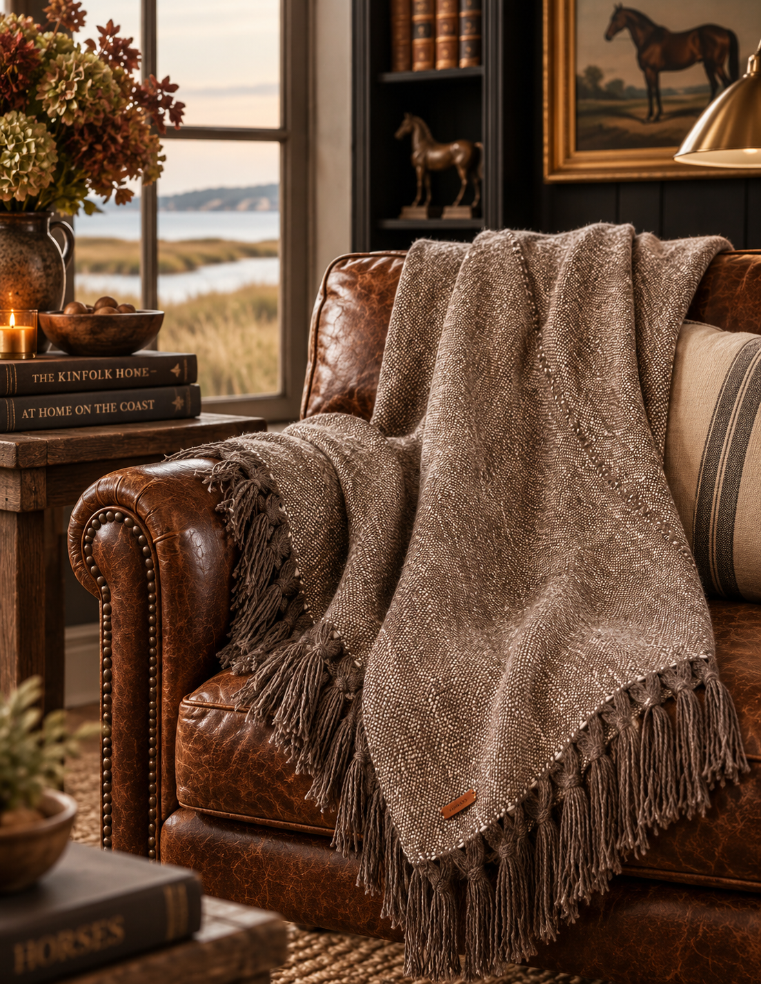 A textured taupe-brown fringed throw blanket is draped over a brown leather armchair in a cozy, rustic living room setting with a window, bookshelf, and warm lighting.