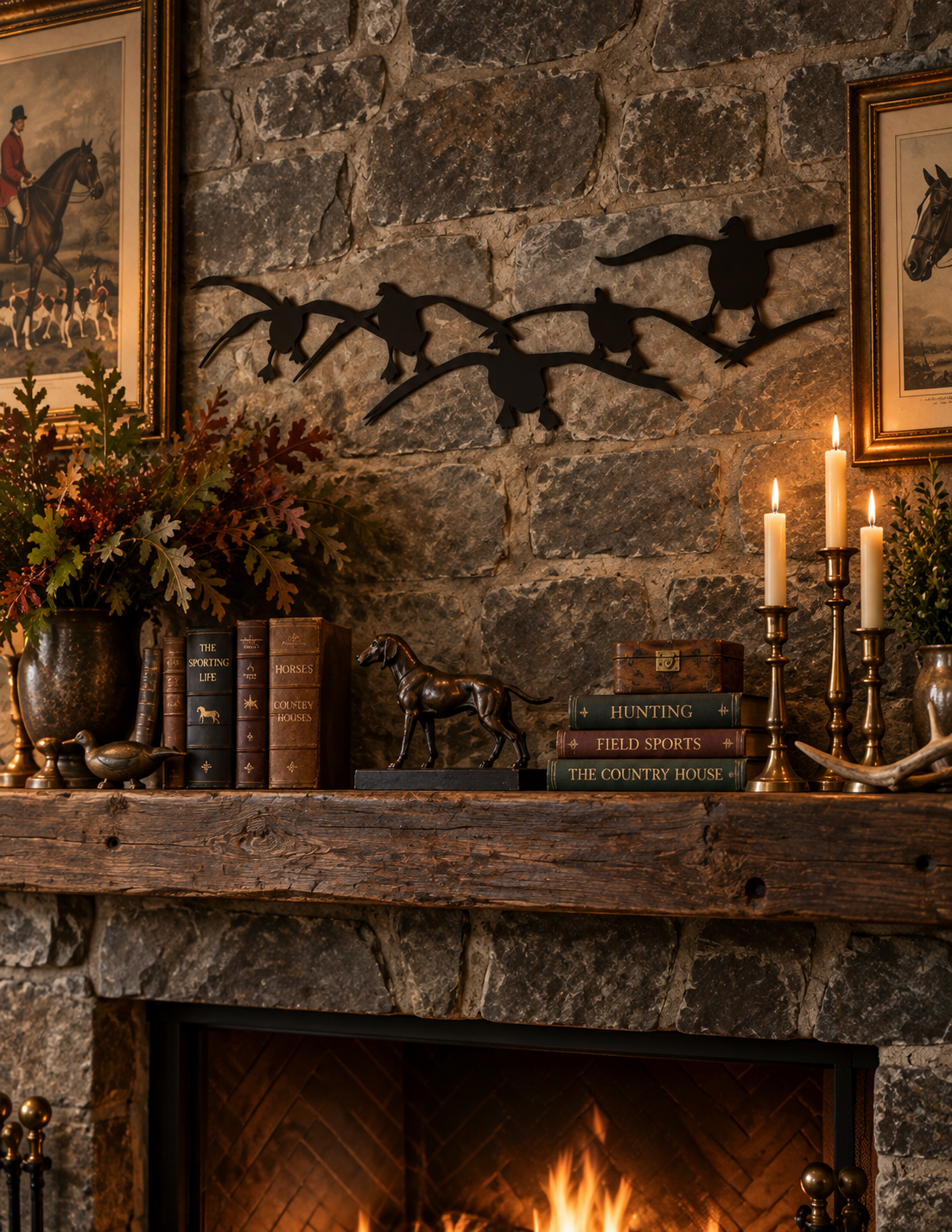 A rustic stone fireplace mantel decorated with framed art, a black metal eagle wall hanging, books, candles, and a small horse figurine, with a warm fire burning below.
