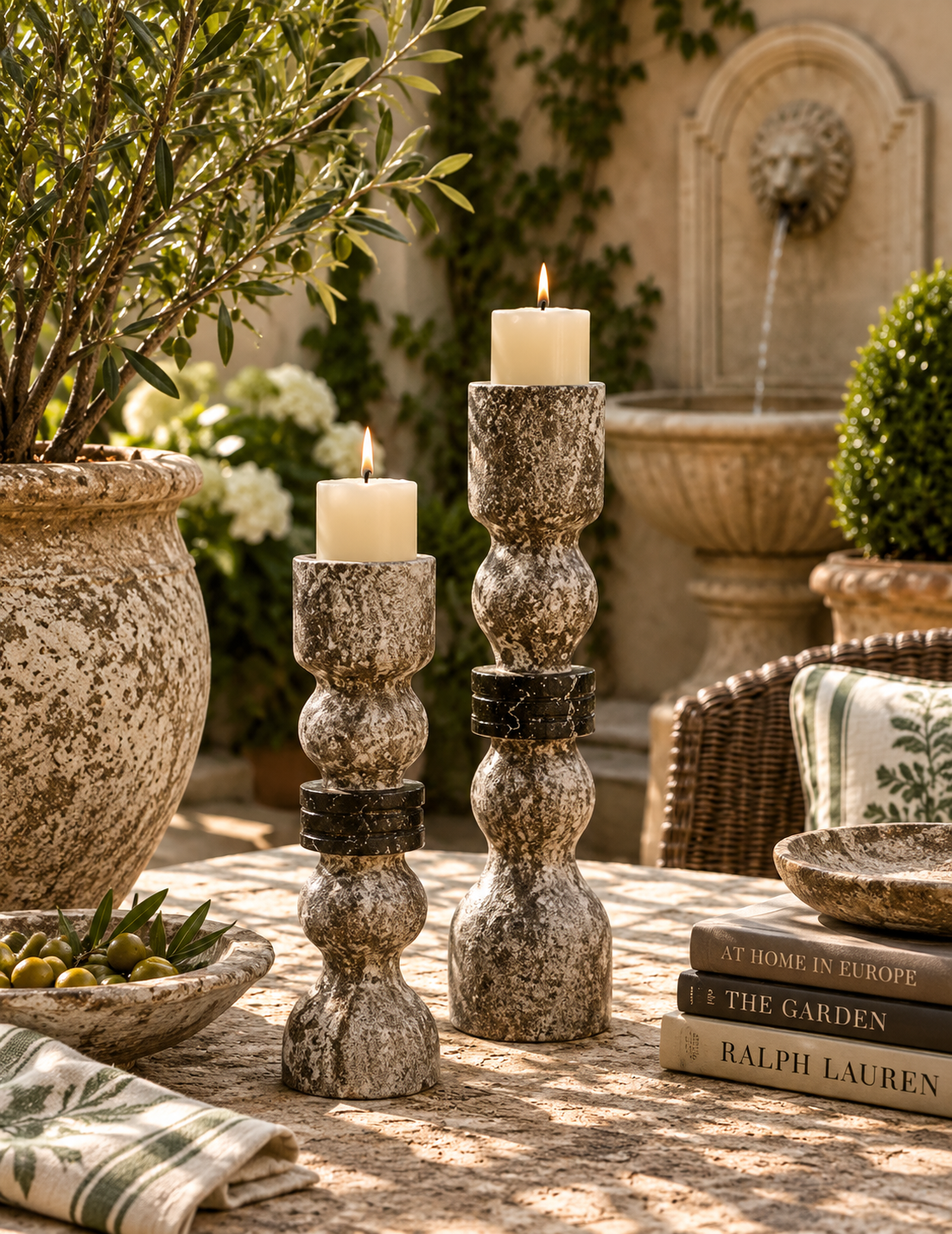 Two distressed stone candlestick holders with lit white candles on an outdoor patio, styled with a bowl of olives, a striped napkin, books, and a fountain in the background.
