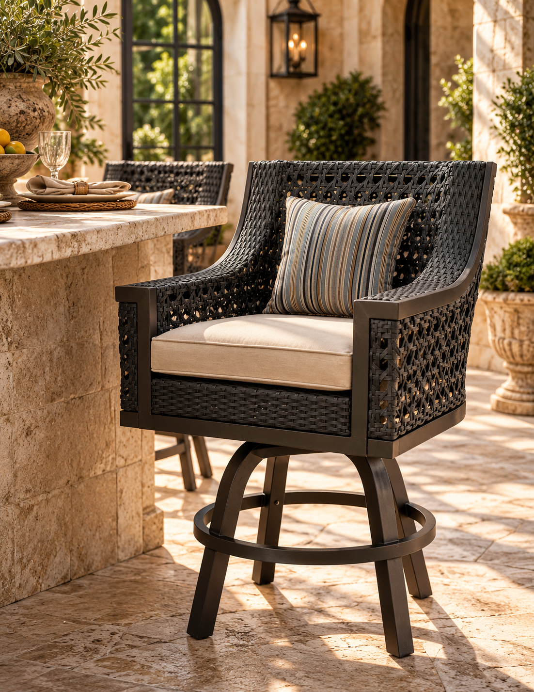 A dark woven wicker bar stool with cream cushions and a striped accent pillow, set on a stone patio next to an outdoor bar counter.