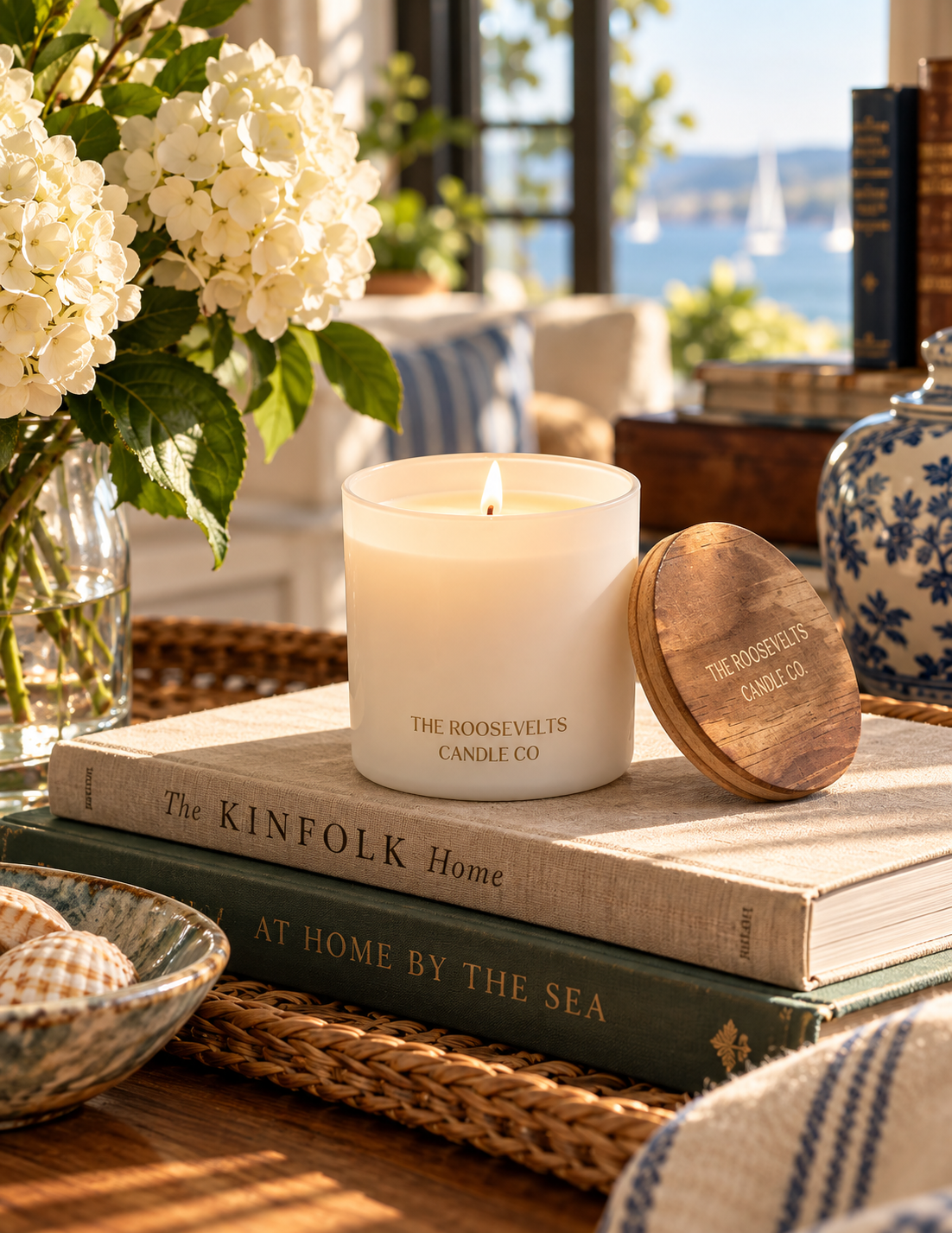 A lit white candle in a clear glass jar sits on stacked books beside a round wooden lid engraved with the brand name, set in a sunlit coastal home interior with flowers, woven textures, and a window view of water and sailboats.