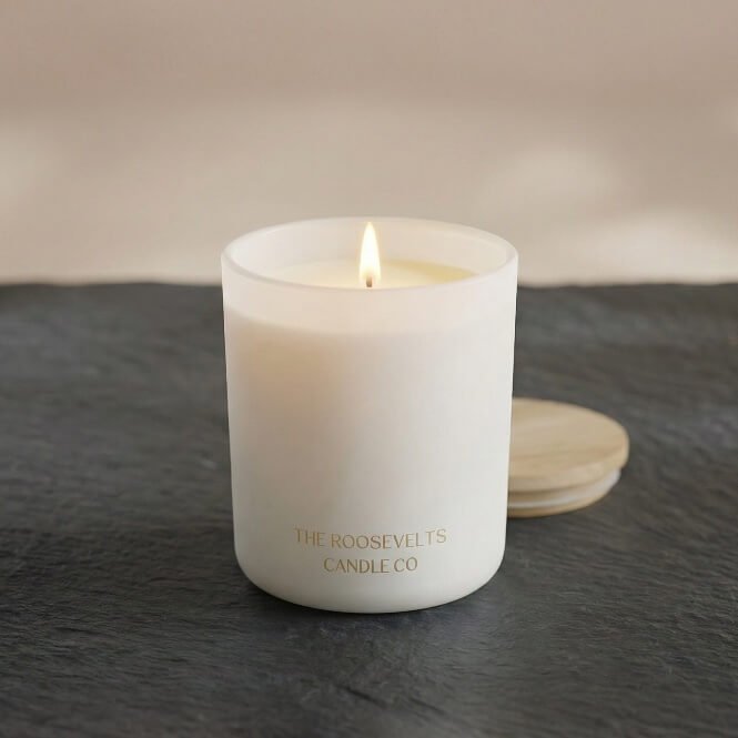 A lit white candle in a frosted glass jar with gold text reading 'THE ROOSEVELTS CANDLE CO' on a dark slate surface, with a light wood lid resting behind it.