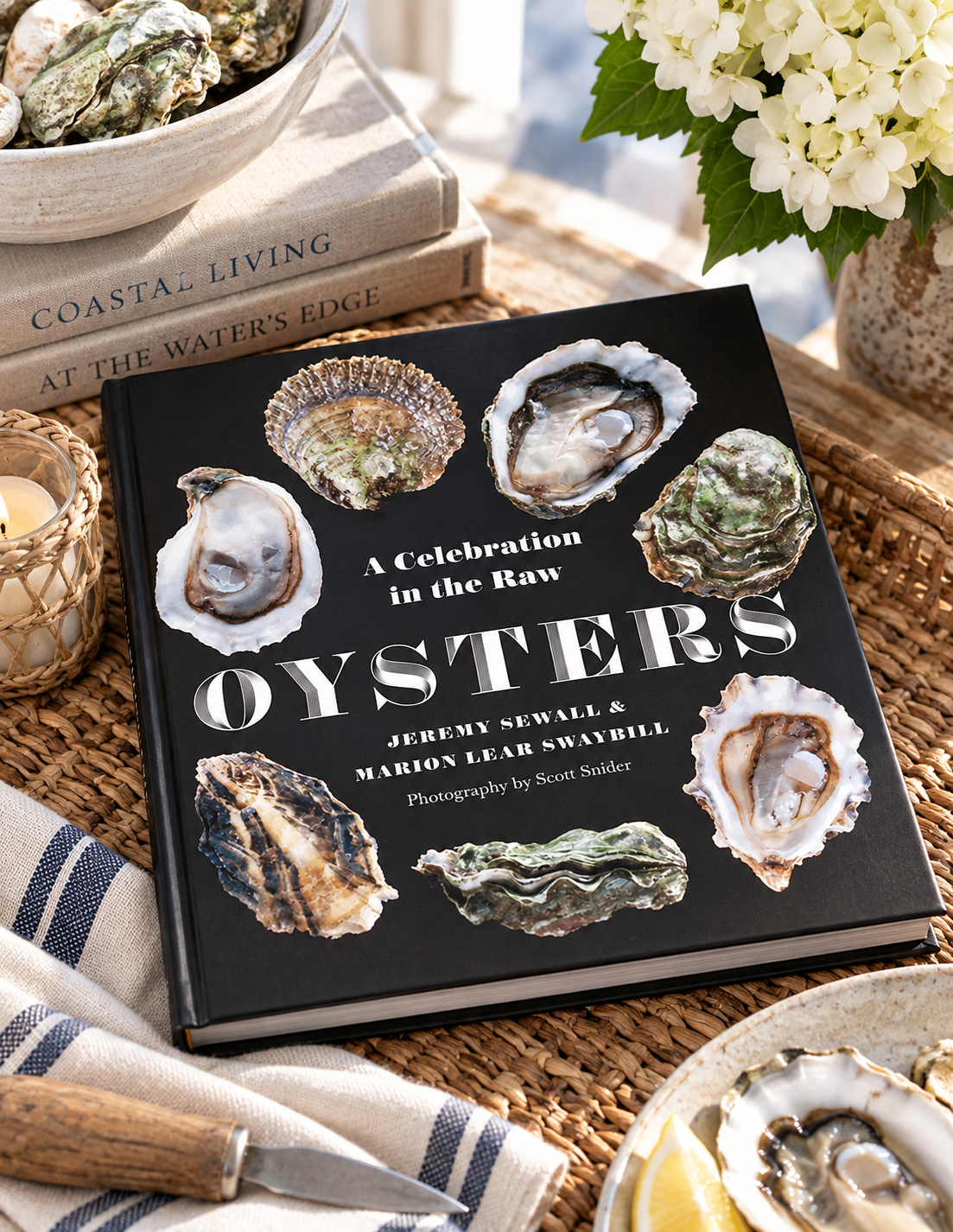A hardcover book titled "Oysters: A Celebration in the Raw" by Jeremy Newall and Marion Lear Swanbill, styled on a woven tray with oysters, a knife, flowers, and other coastal dining props.
